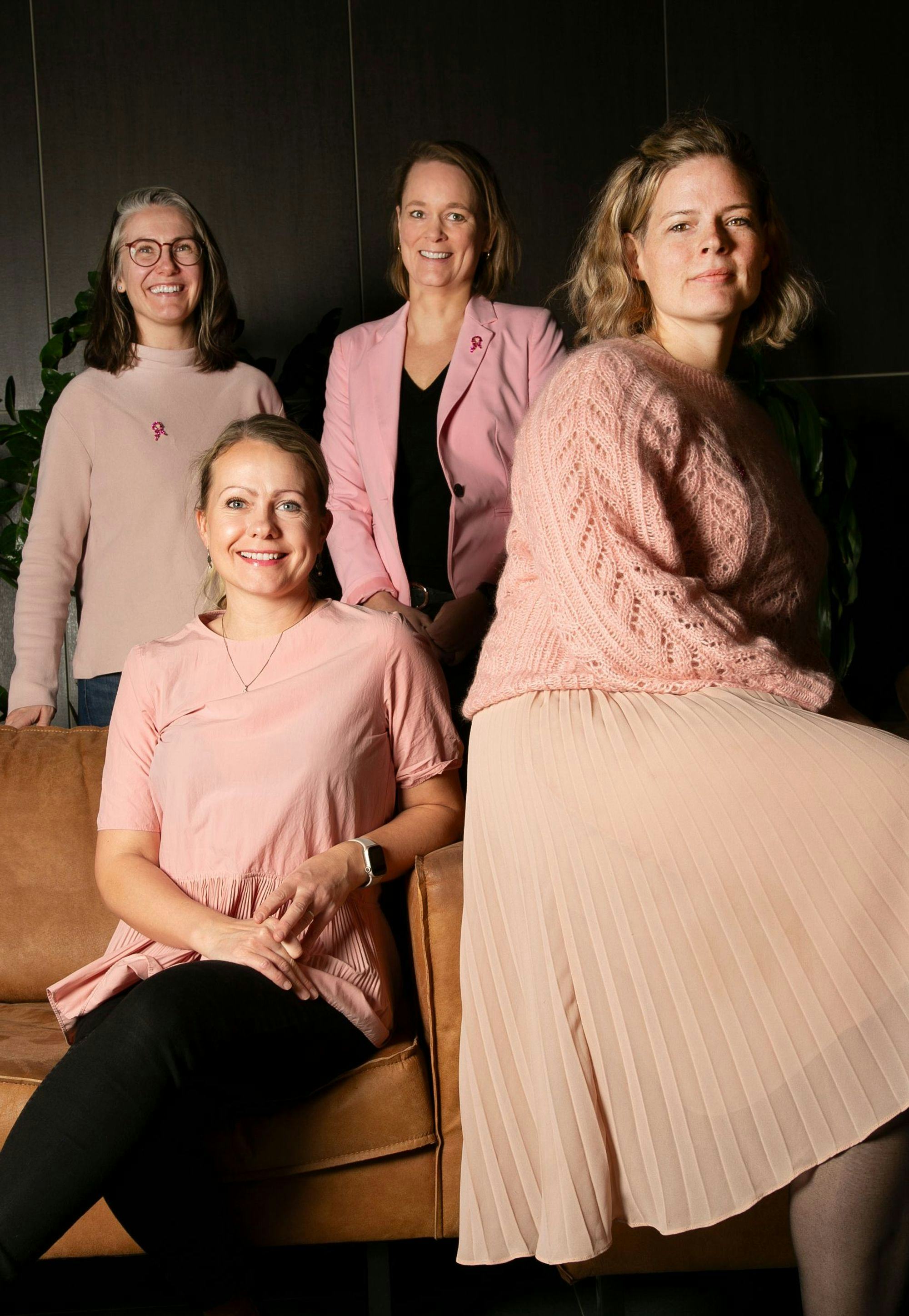 A group of four women, two standing and two seated on a leather couch, all set against a dark interior background with green foliage