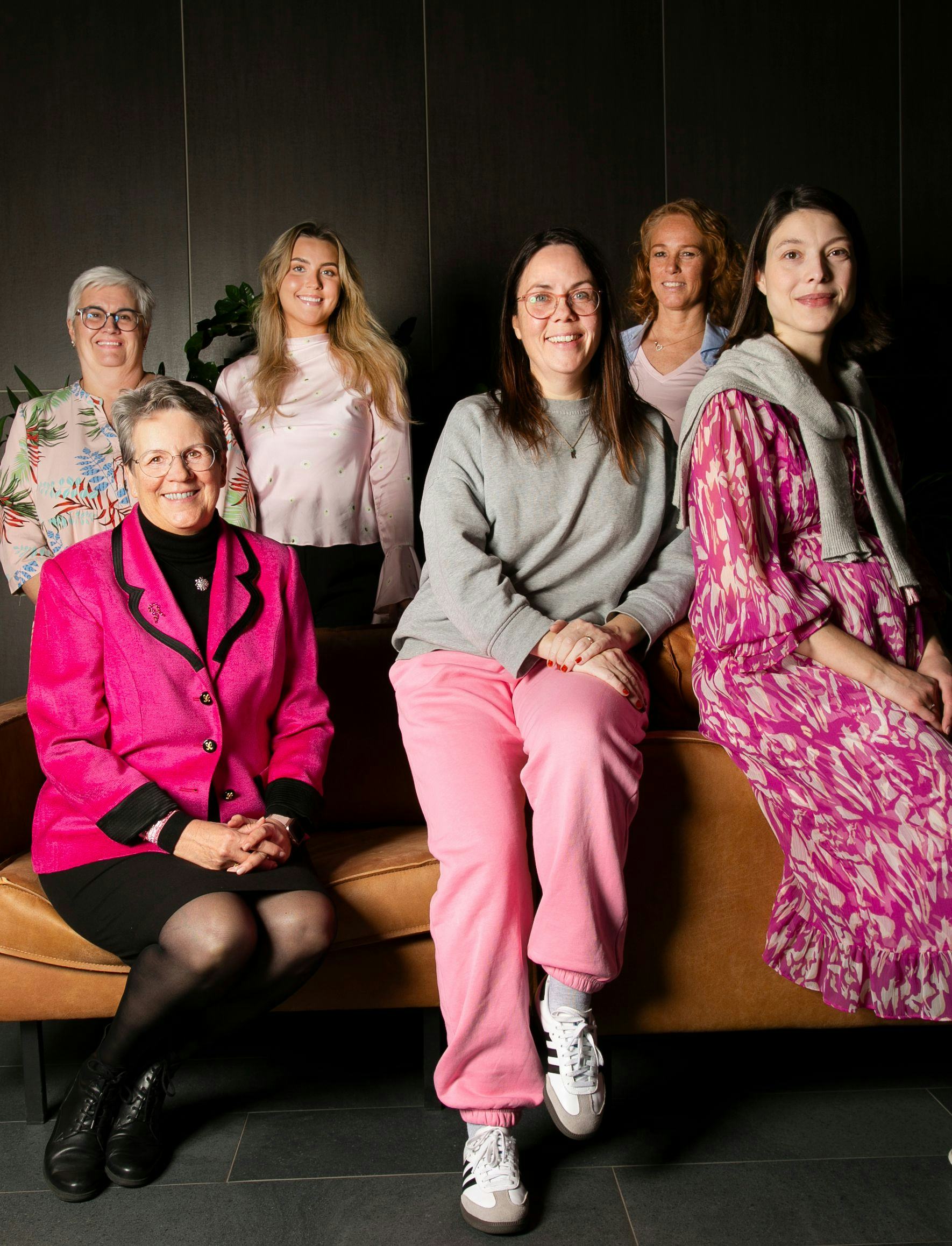 A group of six women posing in an indoor setting with dim lighting. They have varying styles of dresses from casual to formal.
