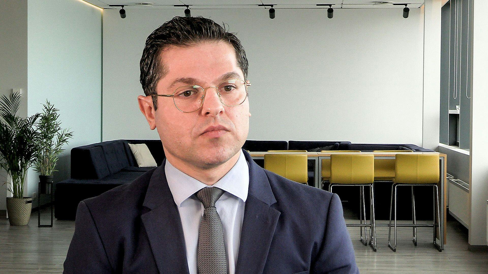 A man in a suit and glasses set against an office background with modern furniture