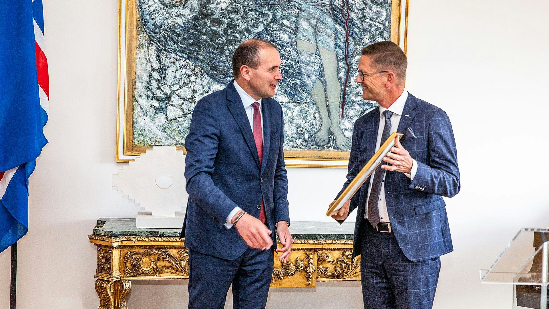 Two men in suits engaging in a conversation, with one holding a frame in a room with an abstract painting in the background