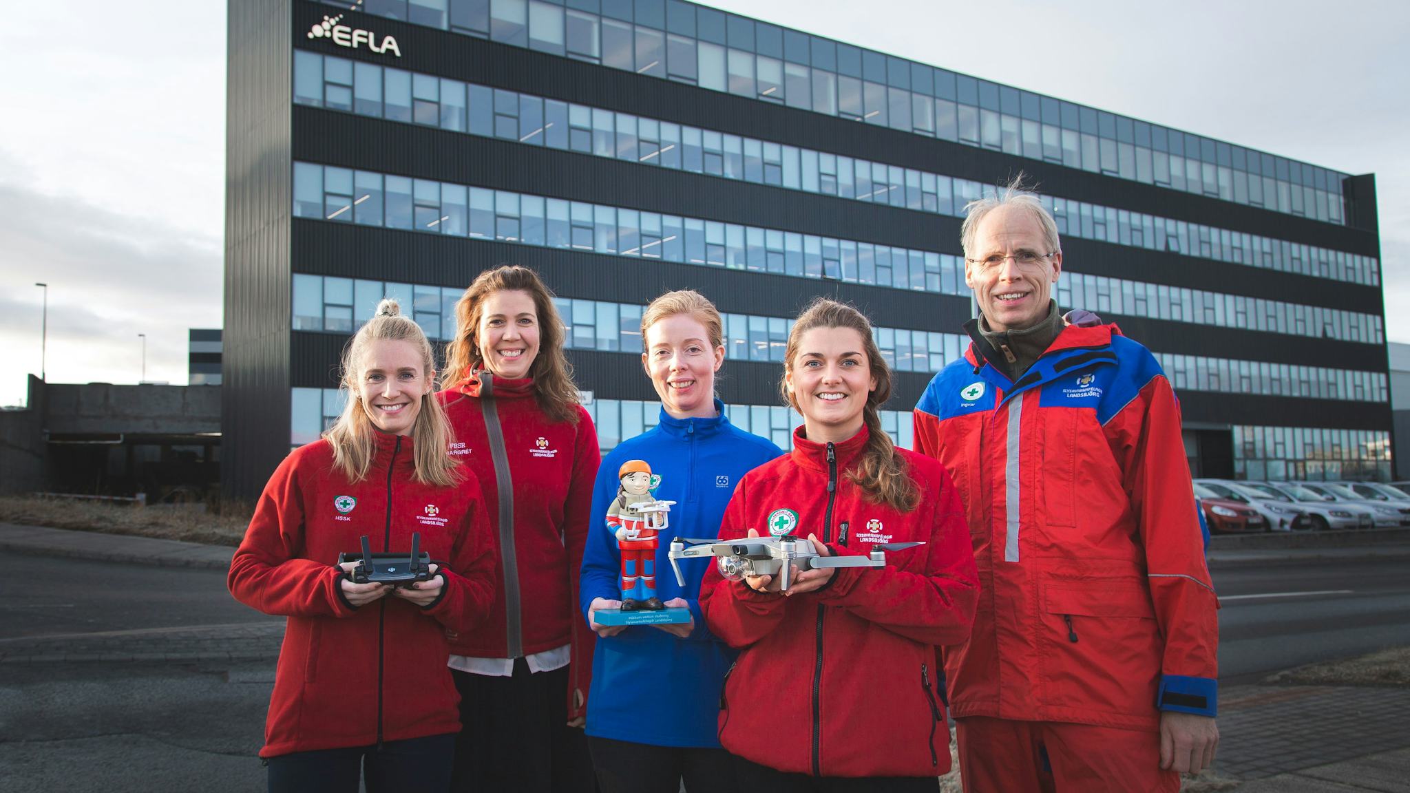 A group of people in red and blue jackets holding drones, in front of a modern building 