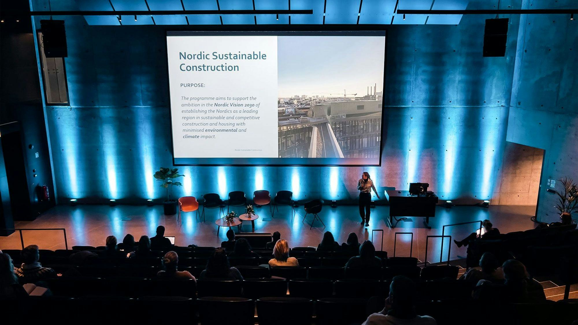 A dark auditorium with a large presentation screen displaying "Nordic Sustainable Construction" material