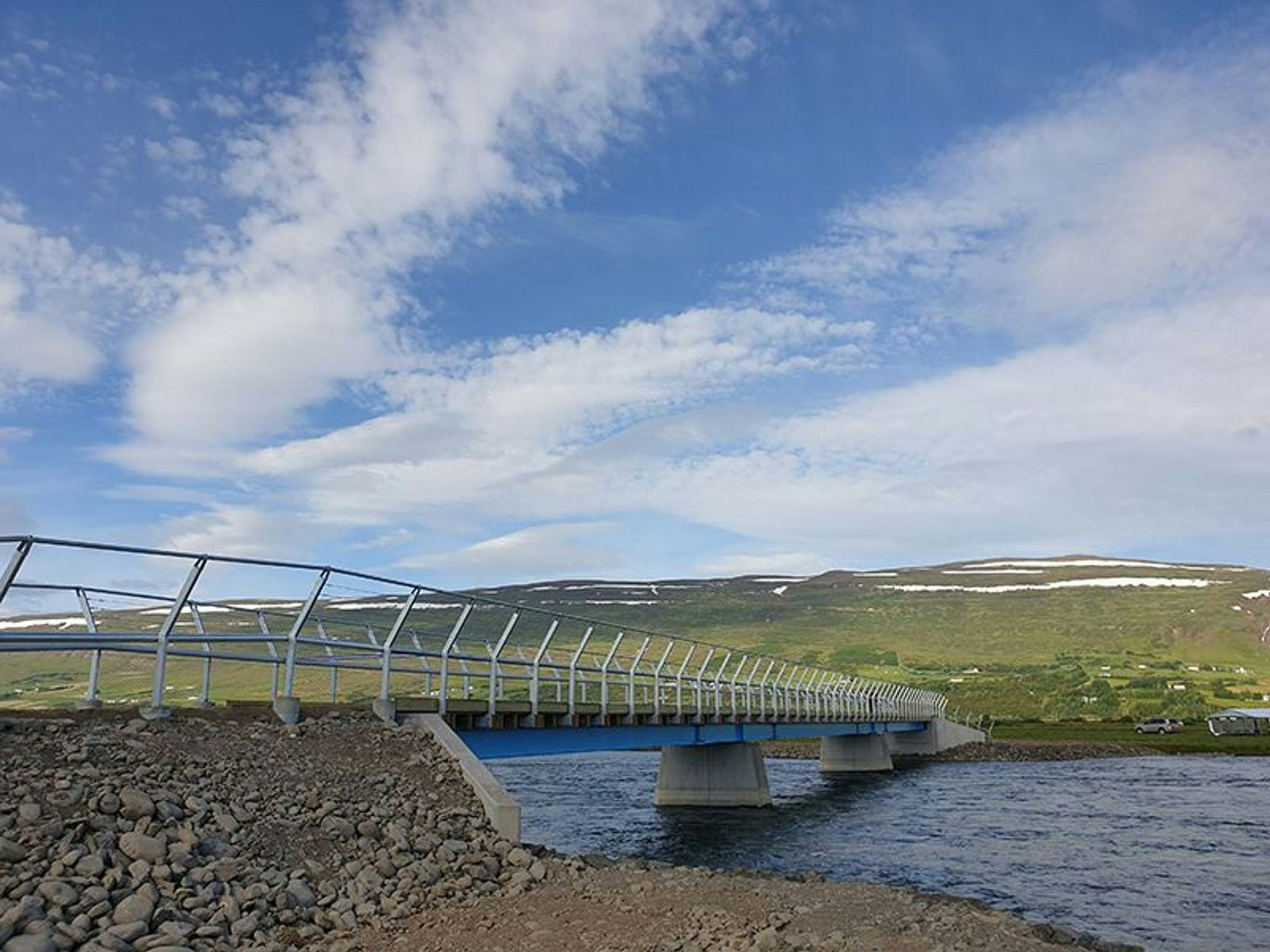 A curving pedestrian bridge with metal railing over a river set against a hilly landscape