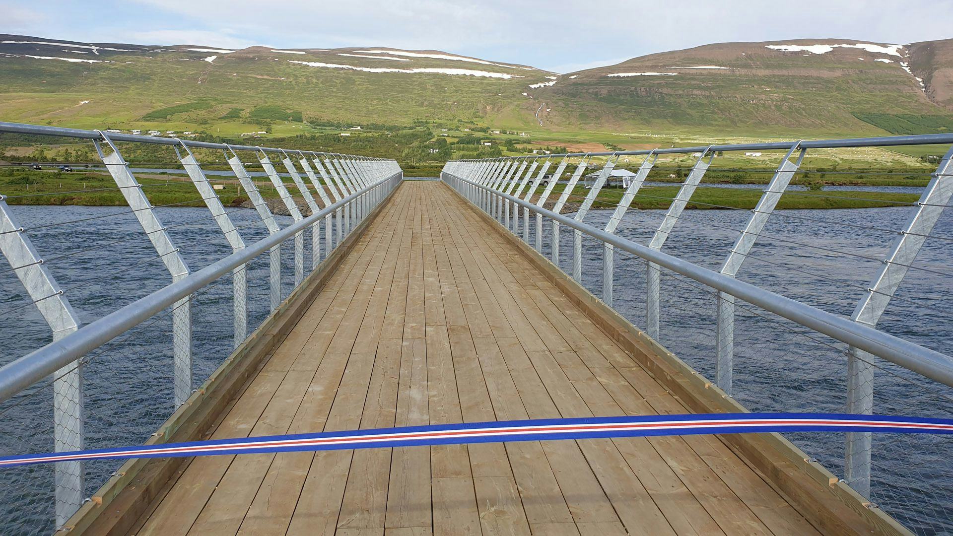 A wooden bridge with metal railings leading towards a hilly landscape