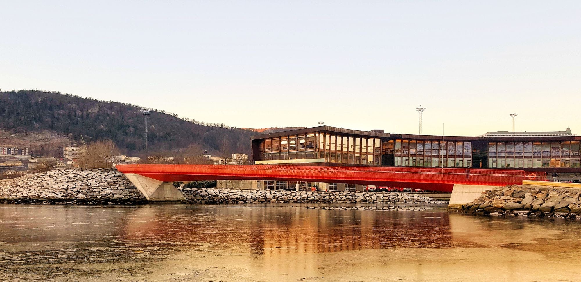 A red bridge spanning a calm river with a modern building and reflective water in the foreground