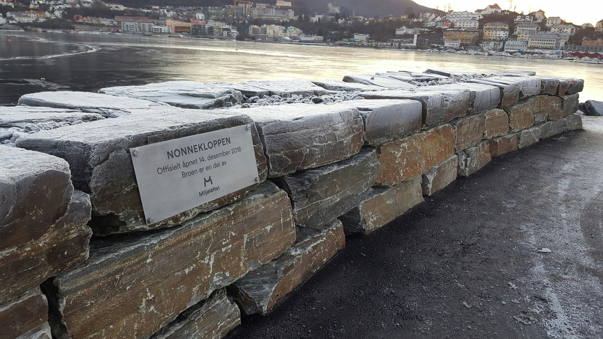 Stone barrier with steel plaque reading "NONNEKLOPPEN" and a view of a city across the water in the background