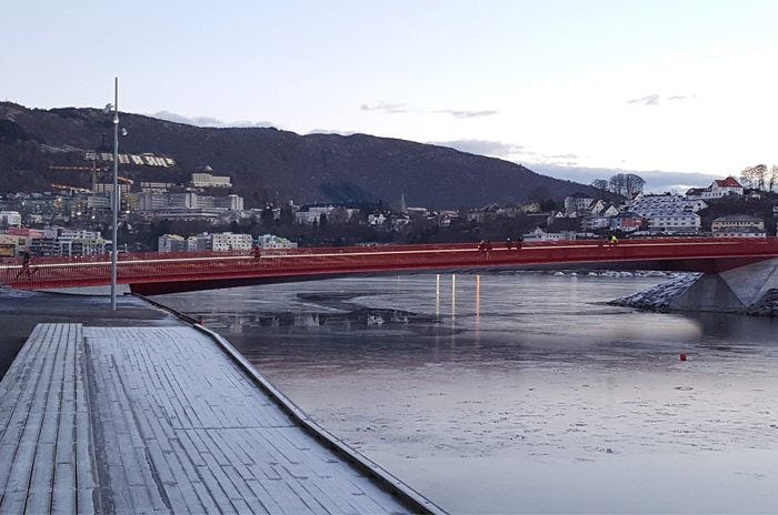 A red bridge against a backdrop of a calm body of water and hilly terrrain