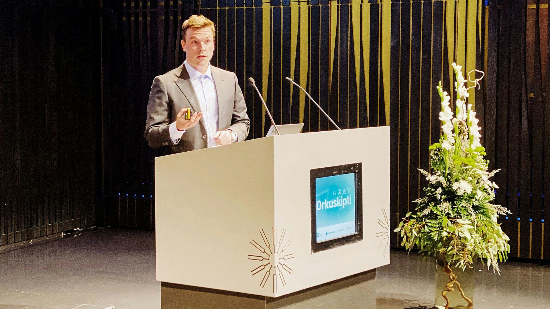 A man giving speech at a podium labelled "orkuskipti" with digital display next to floral arrangement