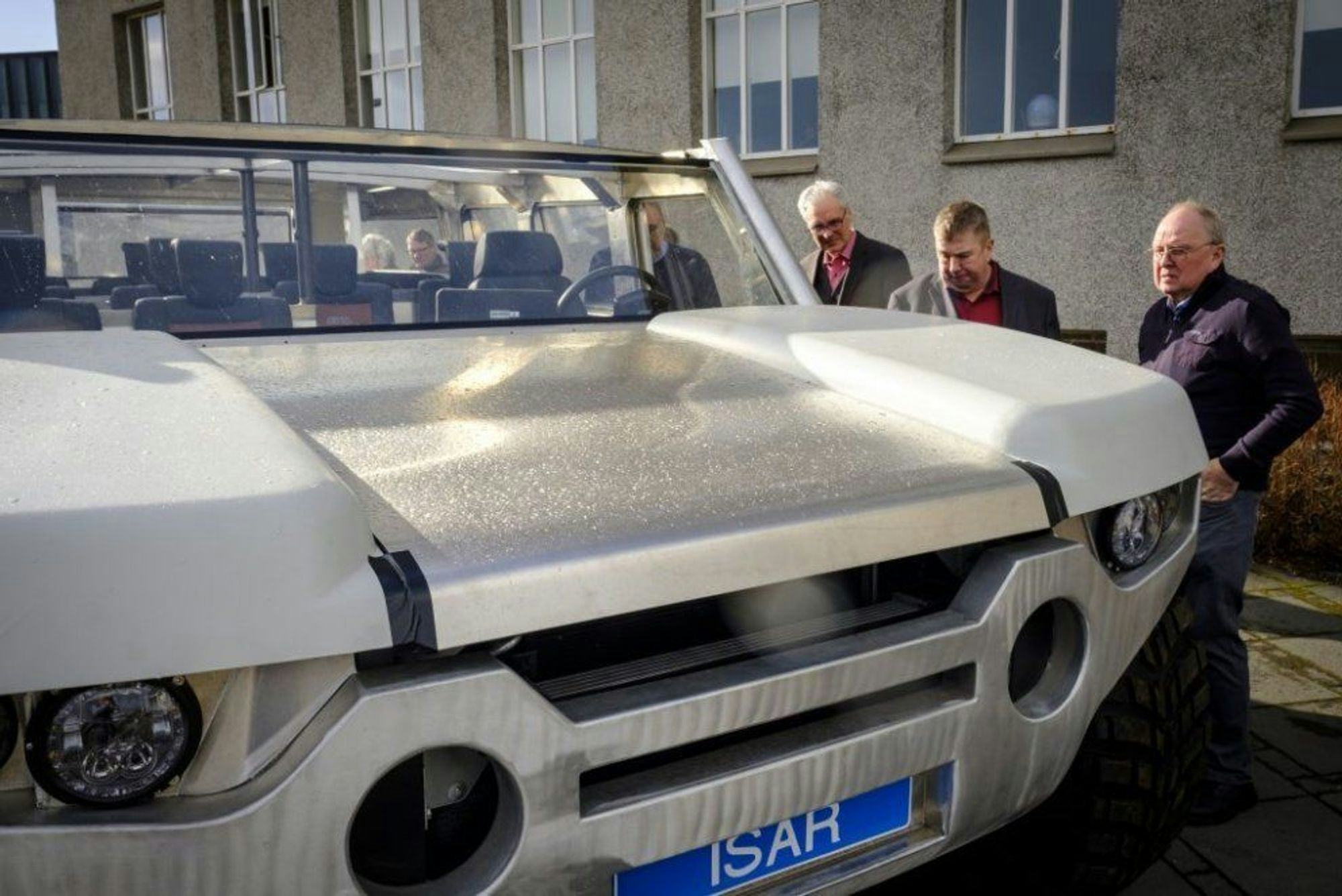 Three men examining a grey car with license plate "ISAR"