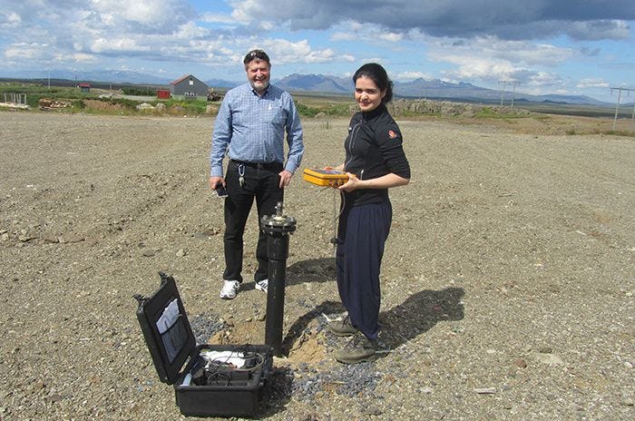A woman holding a yellow box, standing next to a smiling man, testing with some cylindrical object at a landfill site.