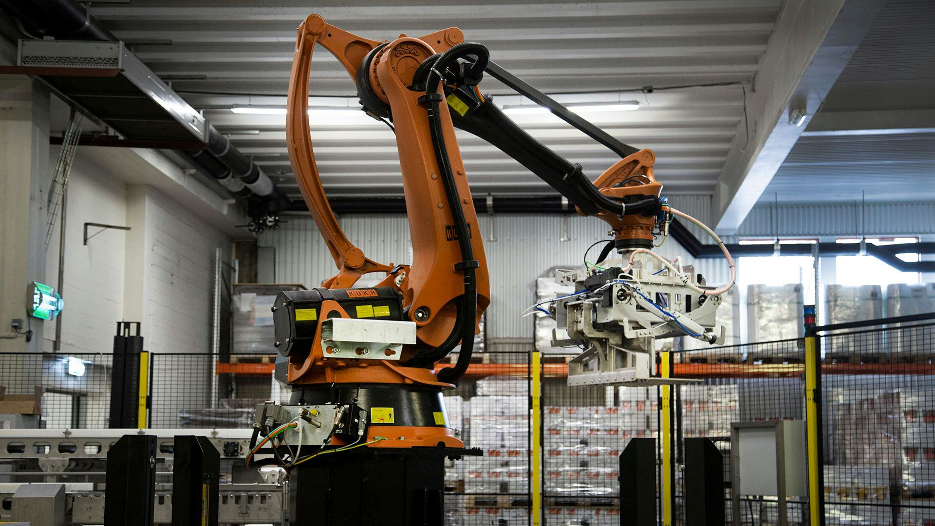 An industrial robot situated in a manufacturing facility with pallets of goods in the background