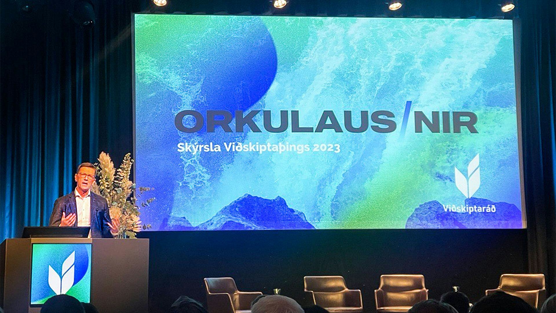 A man is speaking at a lectern with a presentation screen in the background displaying the words "ORKULAUS/N/A"