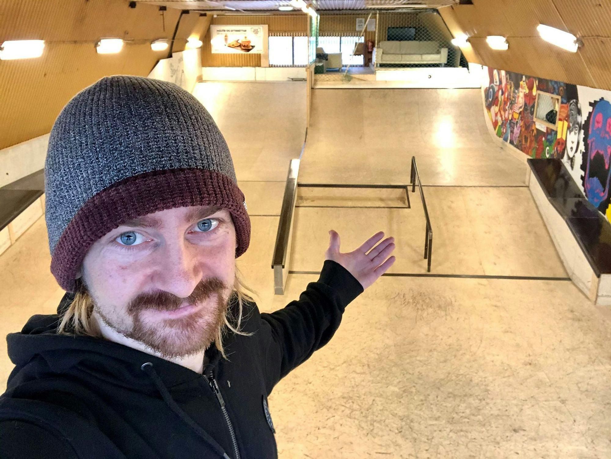 A man with beard and beanie with a hand gesture, in an indoor skatepark in the background