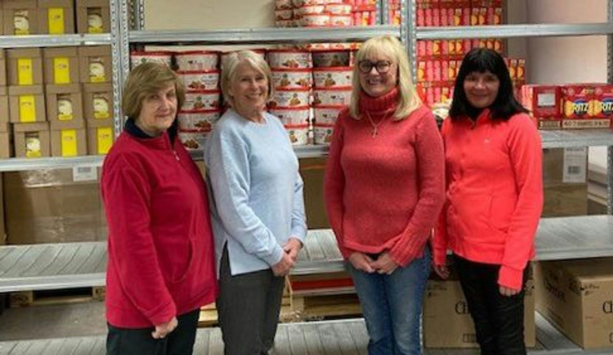 The photo shows four women, three in red and one in blue, smiling and standing together in front of supermarket shelves stocked with products