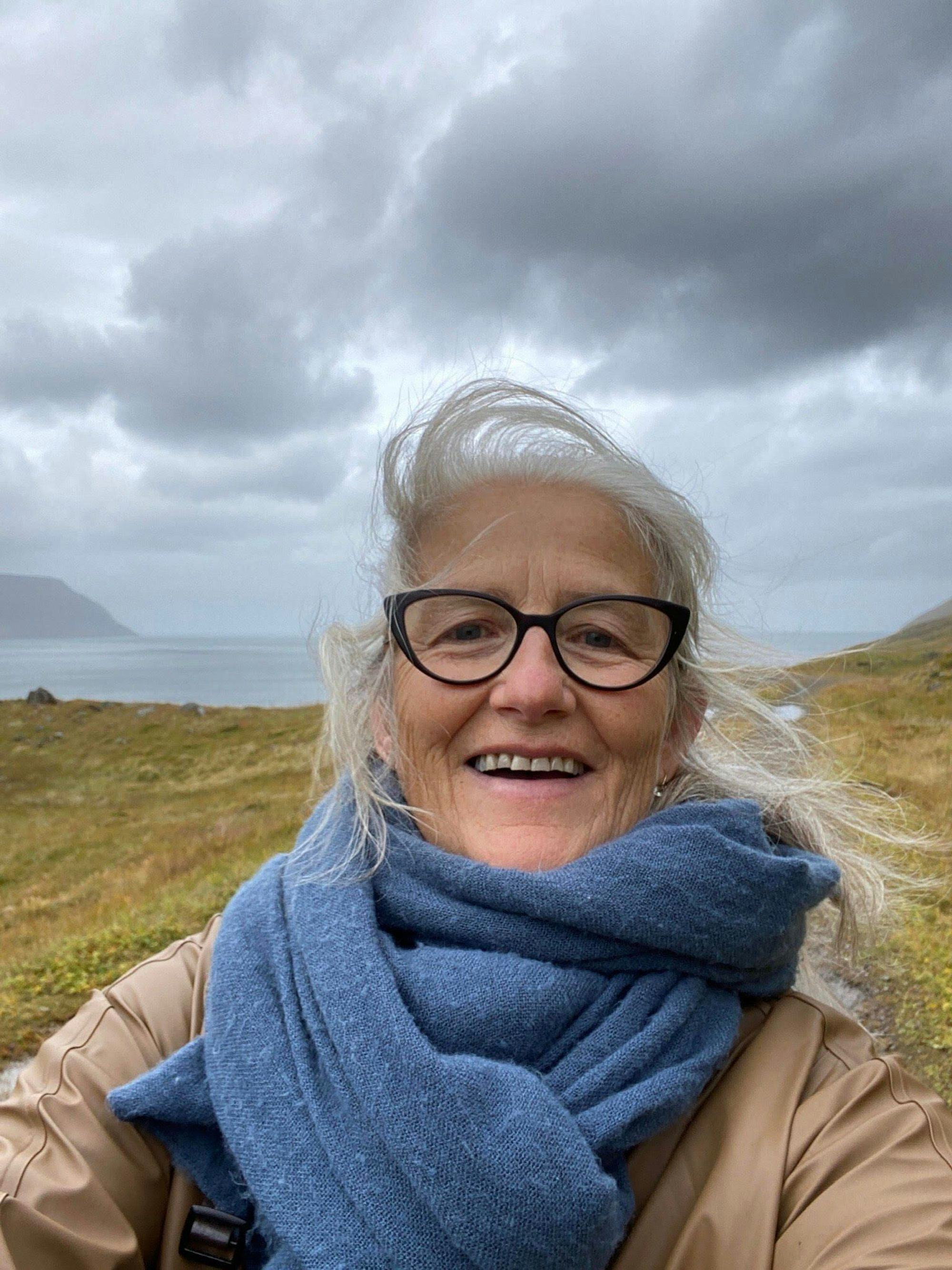 A close-up photo of a smiling woman with gray hair and glasses with a windswept landscape