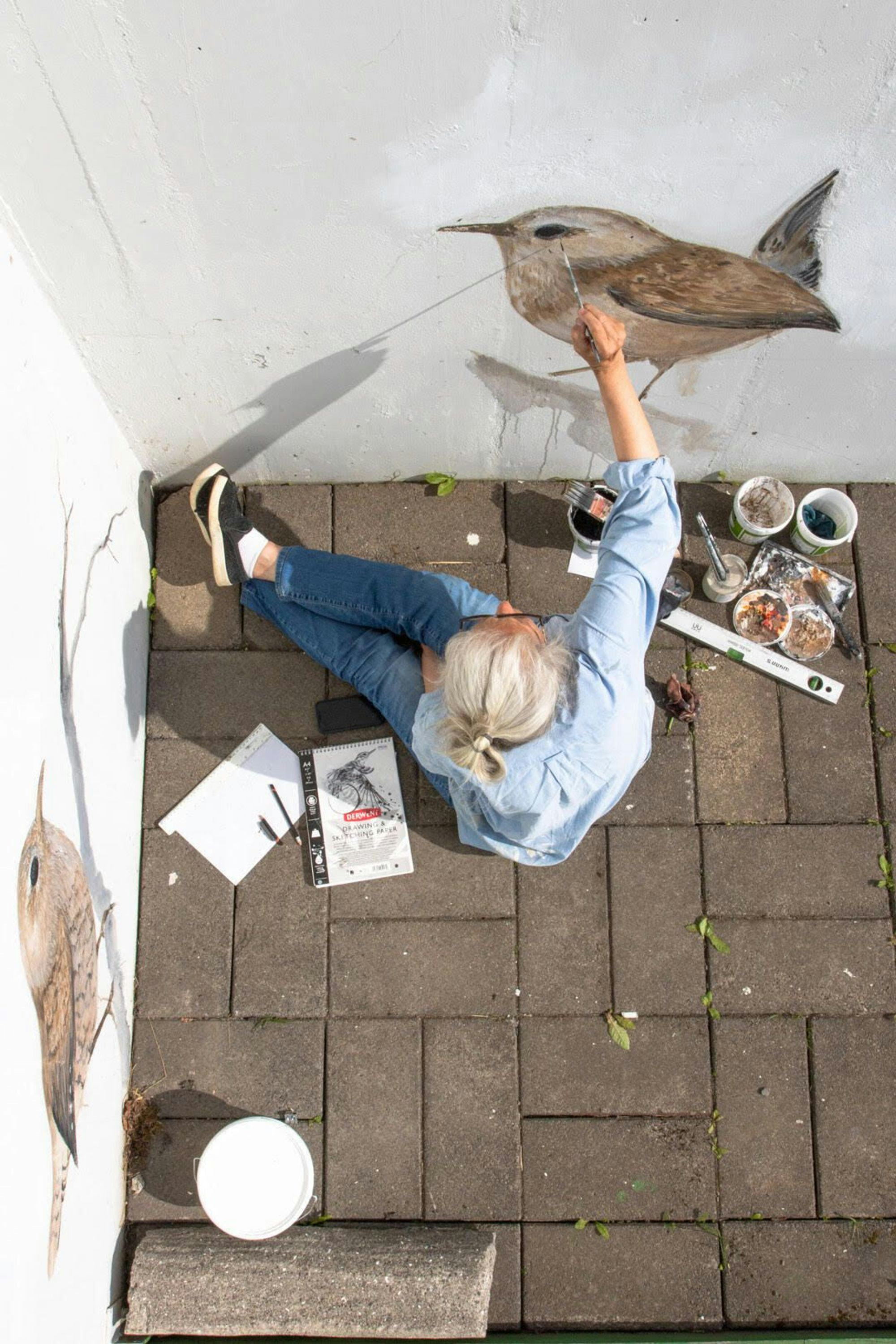 An individual sitting on a brick pavement with art supplies and canvas, viewed from above