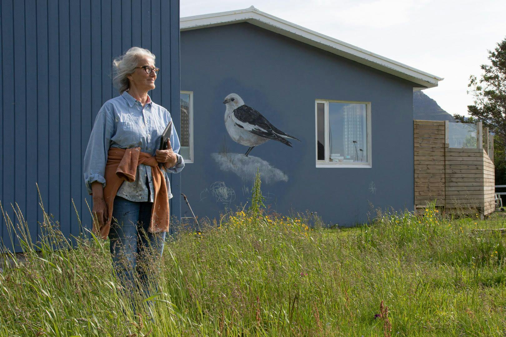 A woman stands in a field of tall grass next to a building with  large mural of a bird on its side