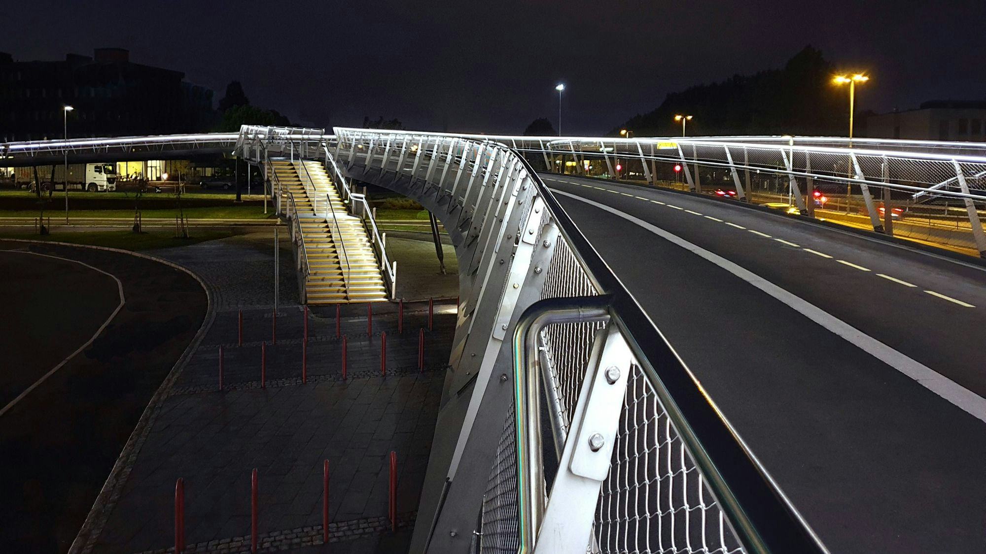 An illuminated pedestrian bridge at night, curving over a road with passing vehicles 