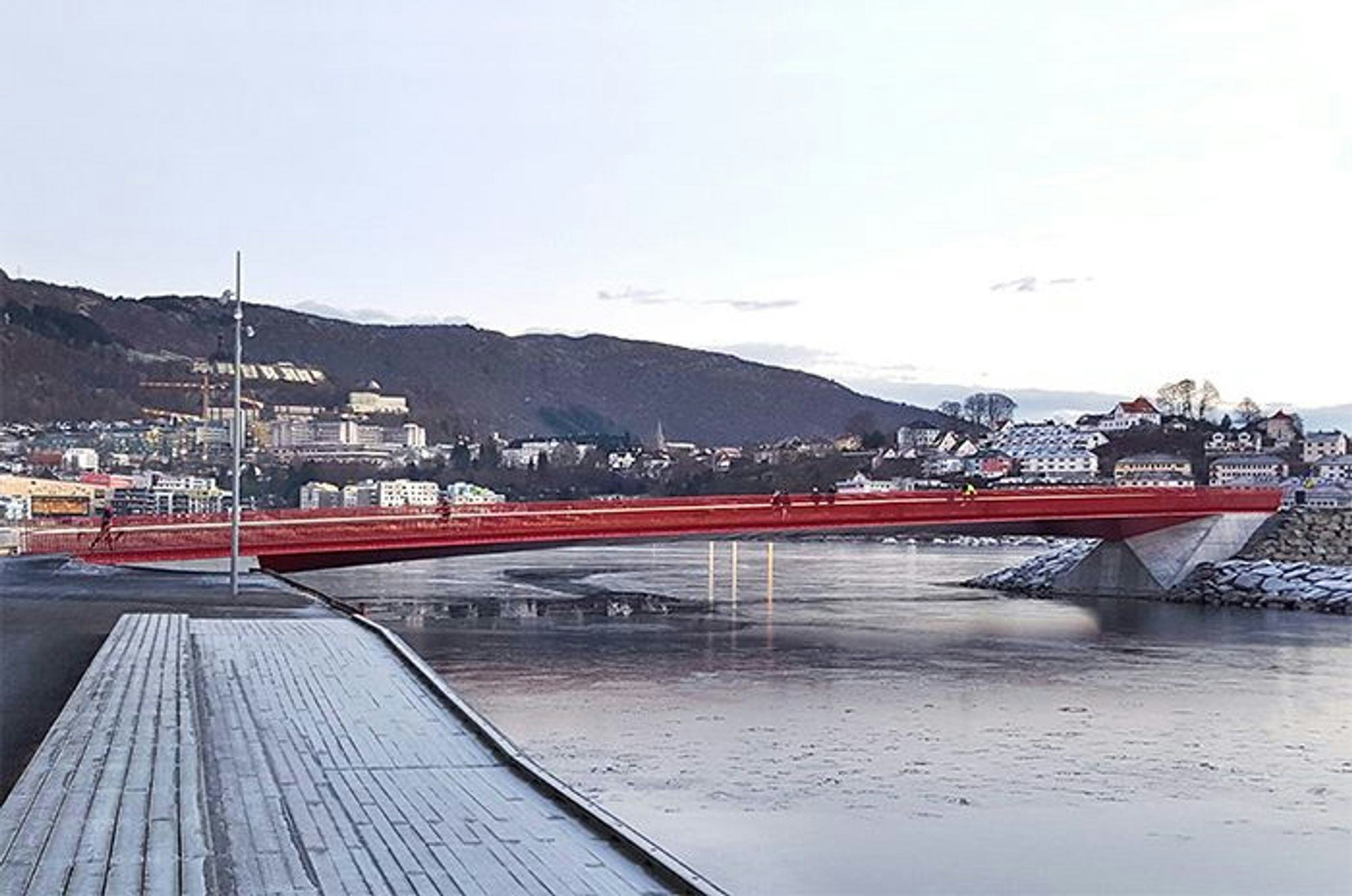 The photo captures a red pedestrian bridge over a calm water with a backdrop of a hilly town under a dusky sky