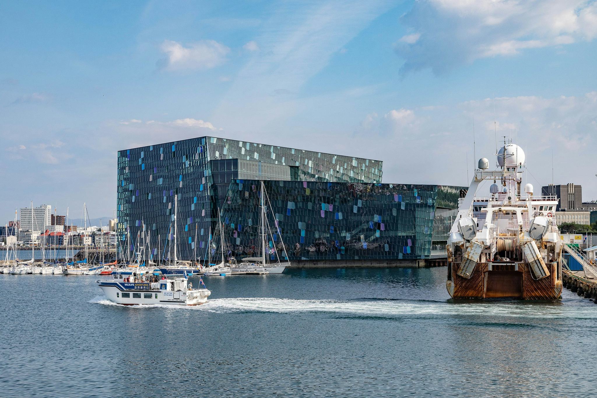A modern building with unique patterned facade by the waterfront alongside a variety of boats including a yacht and a large ship