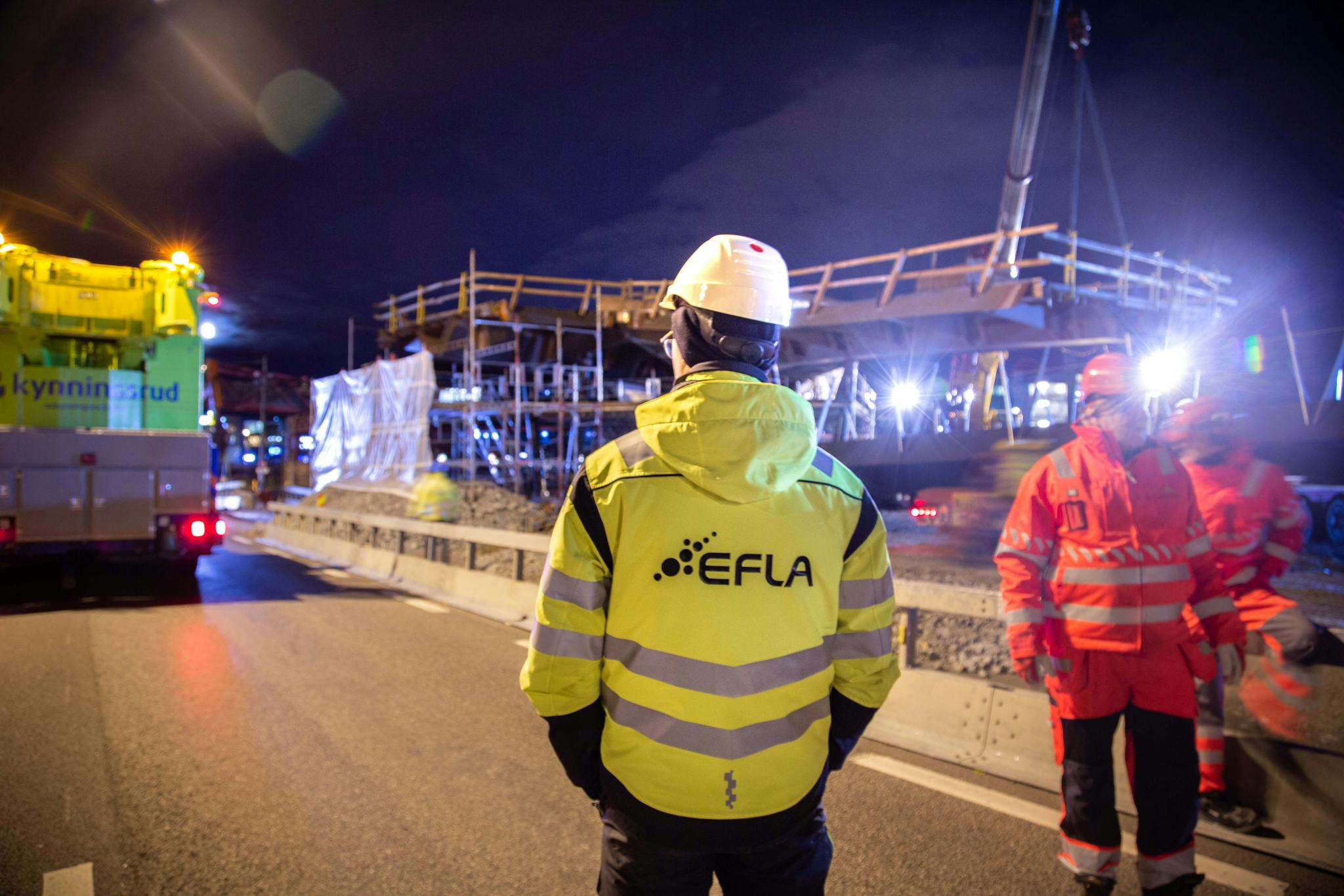 Three men wearing high visibility jacket at a brightly lit construction site at night
