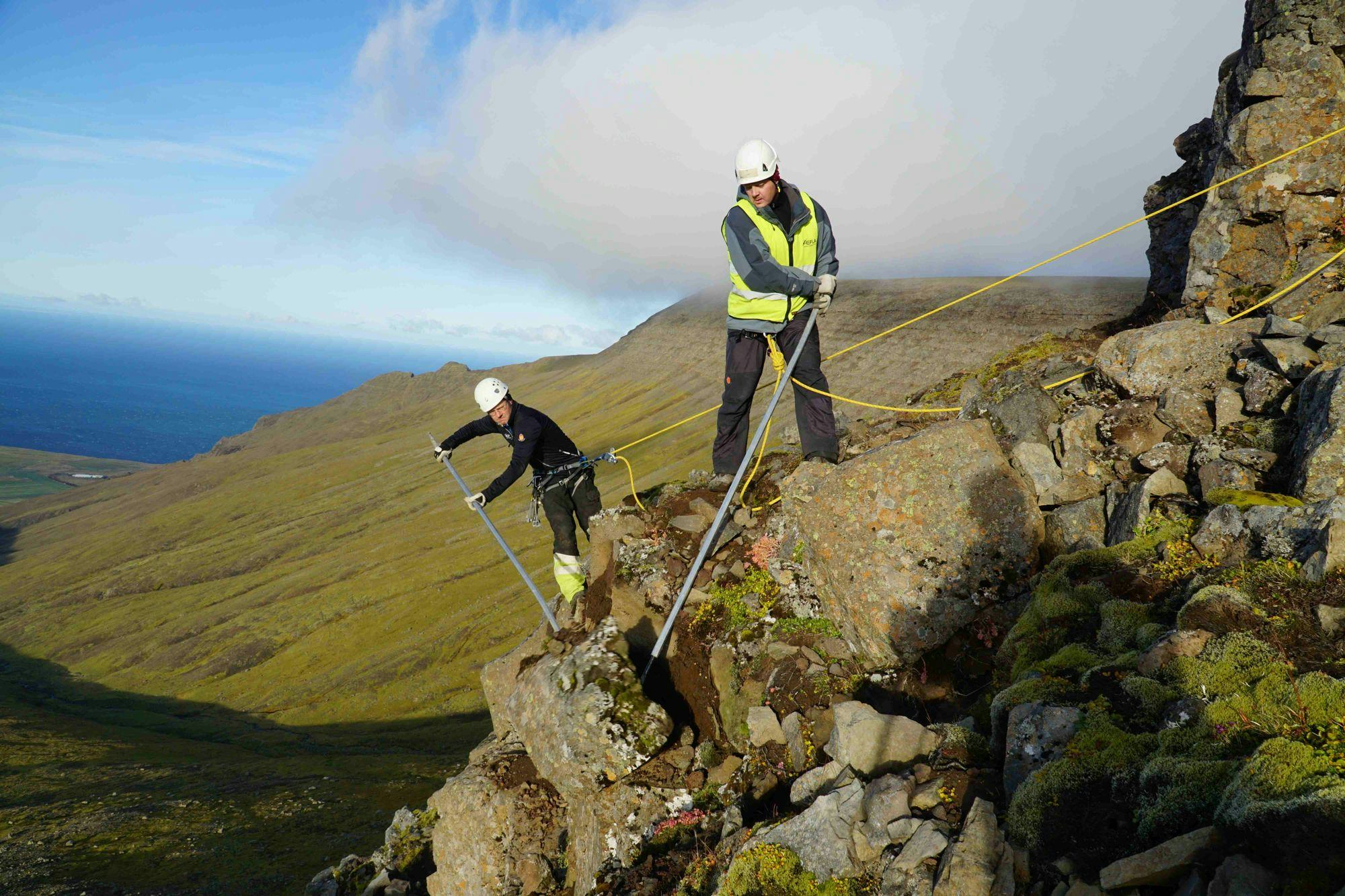 Two individuals wearing high visibility jacket, working on a rocky hillside with rope