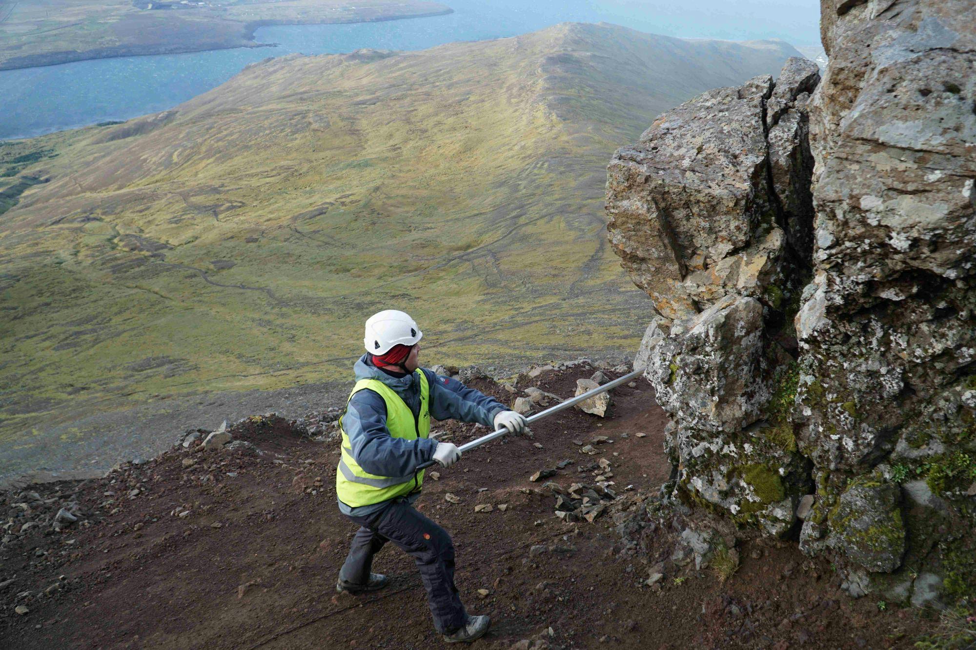 An individual wearing high visibility jacket, working on a rocky hillside with a stick
