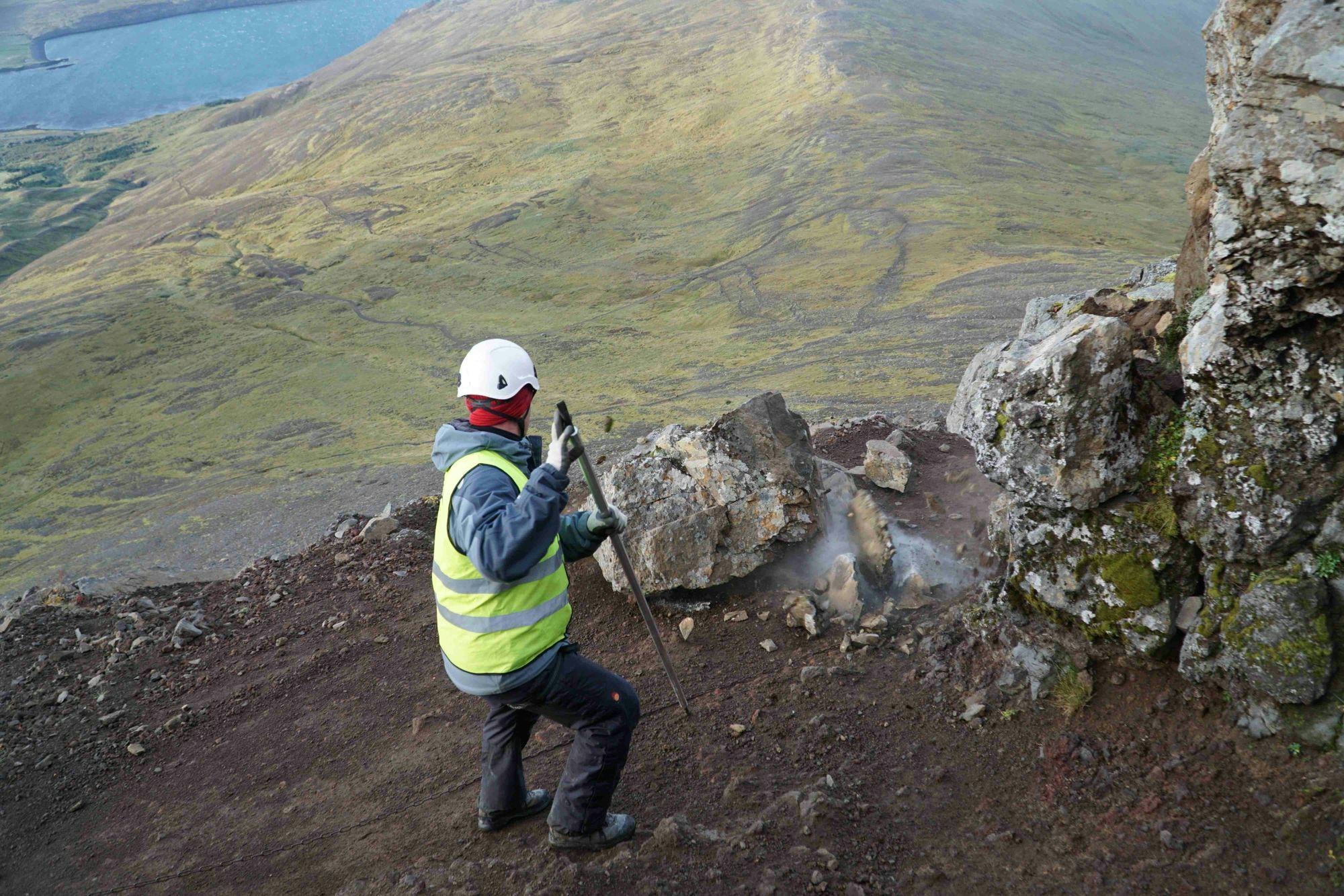 An individual wearing high visibility jacket, working on a rocky hillside with a stick