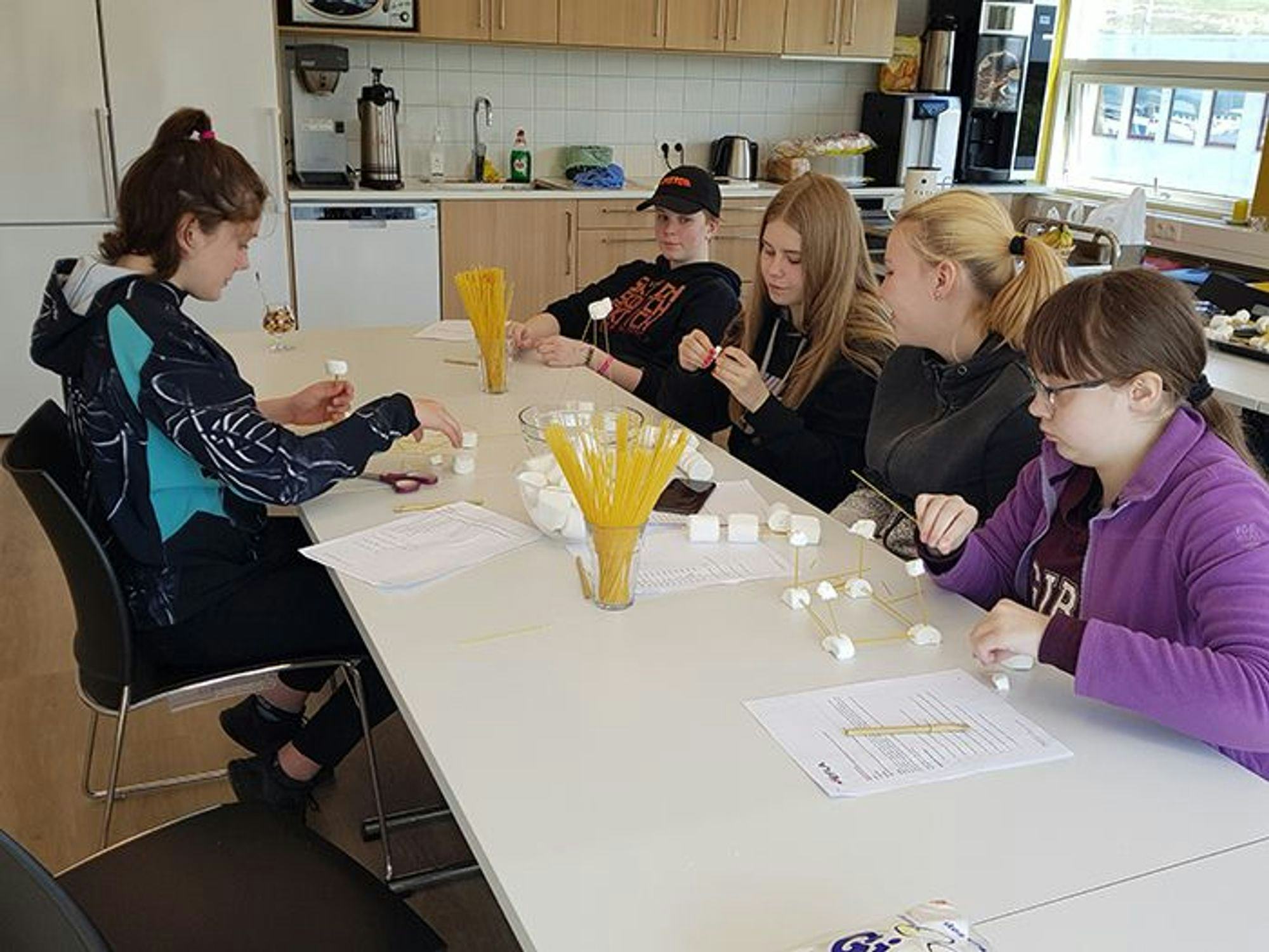 Five young people seated at a long white table, with spaghetti and marshmallow on it