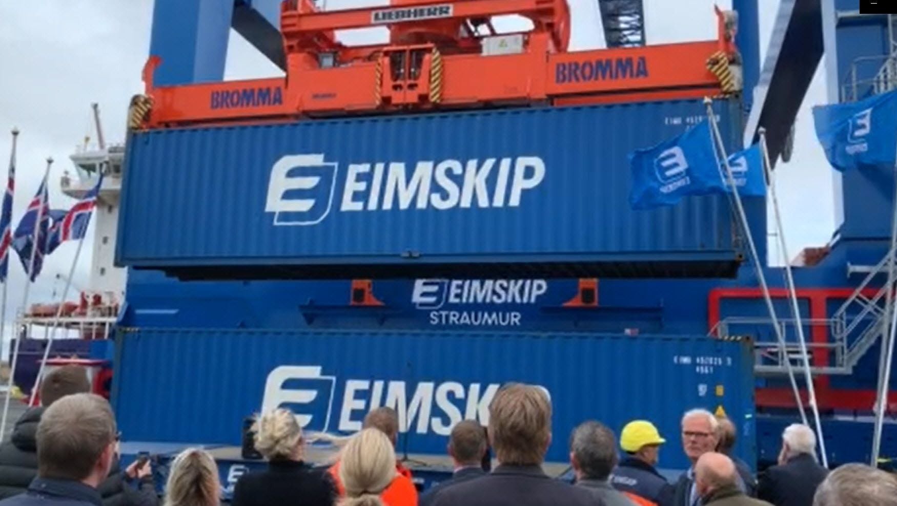 People observing a large piece of orange machinery on top of Blue big shipping containers
