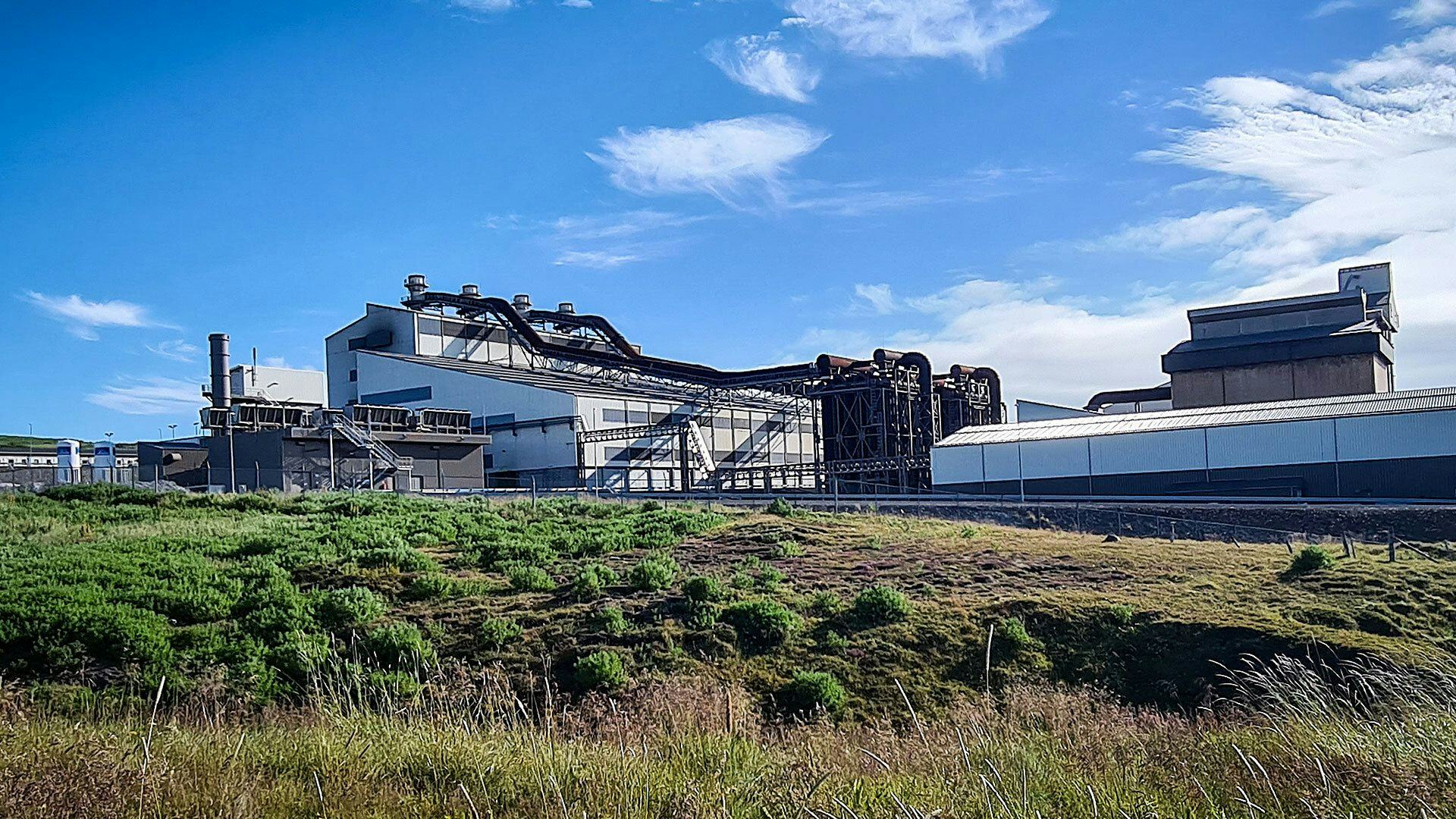 The image shows an industrial facility with large plant and processing facility set against a clear sky with some cloud cover