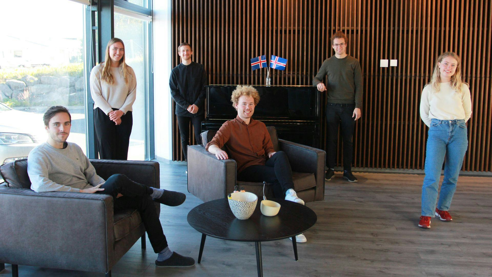 A group of young individuals posing in a modern interior space with a piano and Icelandic flags in the background