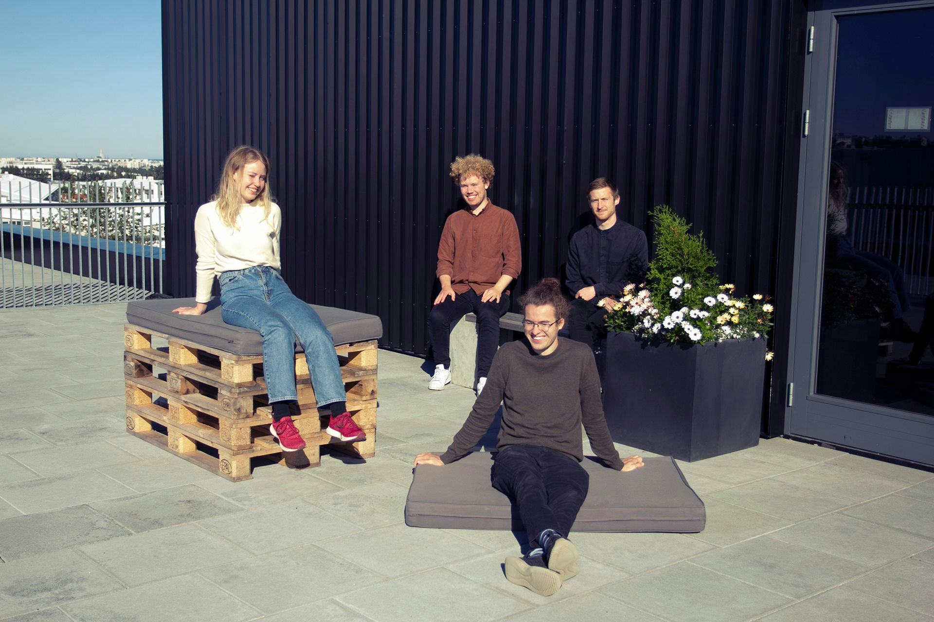 Four individuals relaxing on a sunny rooftop with seating made from pallets and cushions