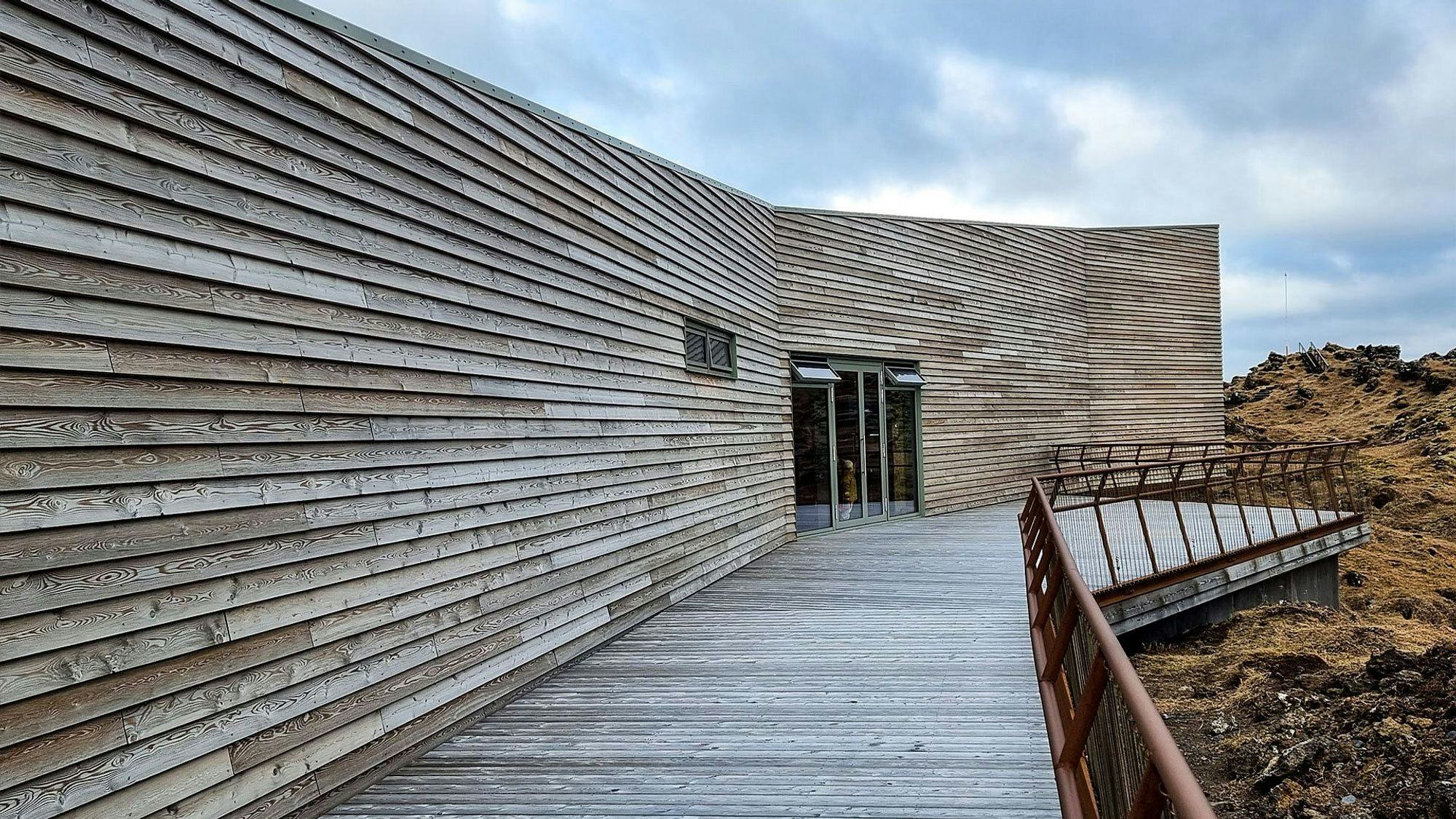 A wooden walkaway leading to a modern building with horizontal slat siding in a natural setting