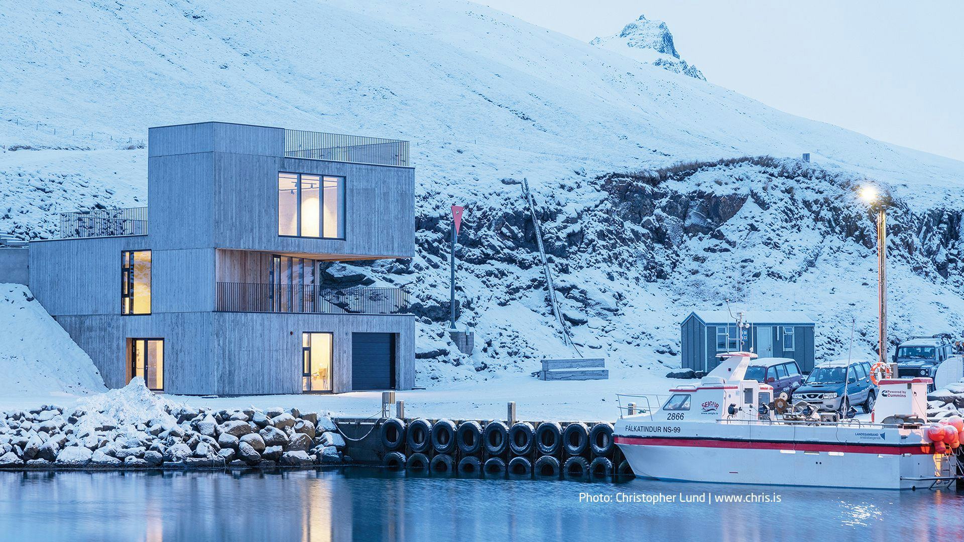 A modern architectural building by the water with a backdrop of a snowy mountain landscape and a boat docked at the pier