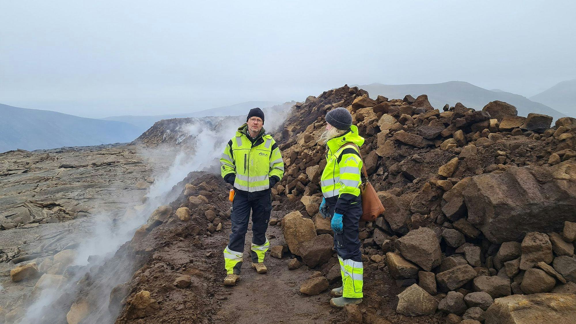 Two individuals in high visibility jackets are standing on a rocky terrain with steam rising in the background