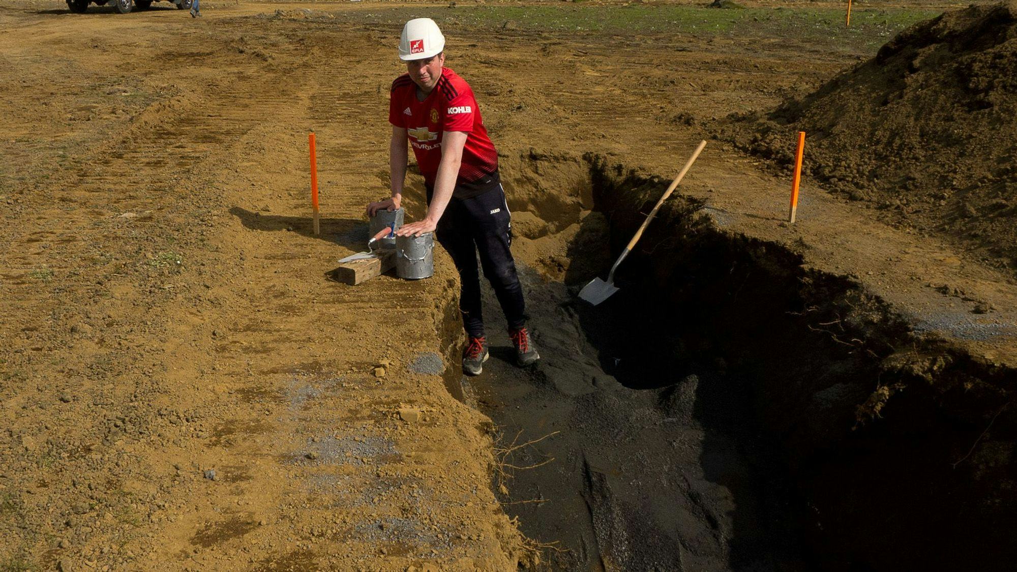 a man wearing a red shirt and a white hard hat in an excavation area holding a bucket 