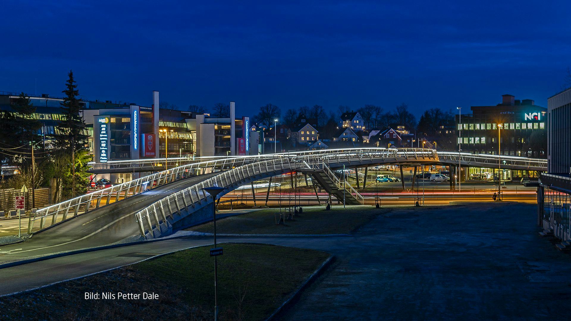 A curving pedestrian bridge at dusk with the lights of a city in the background