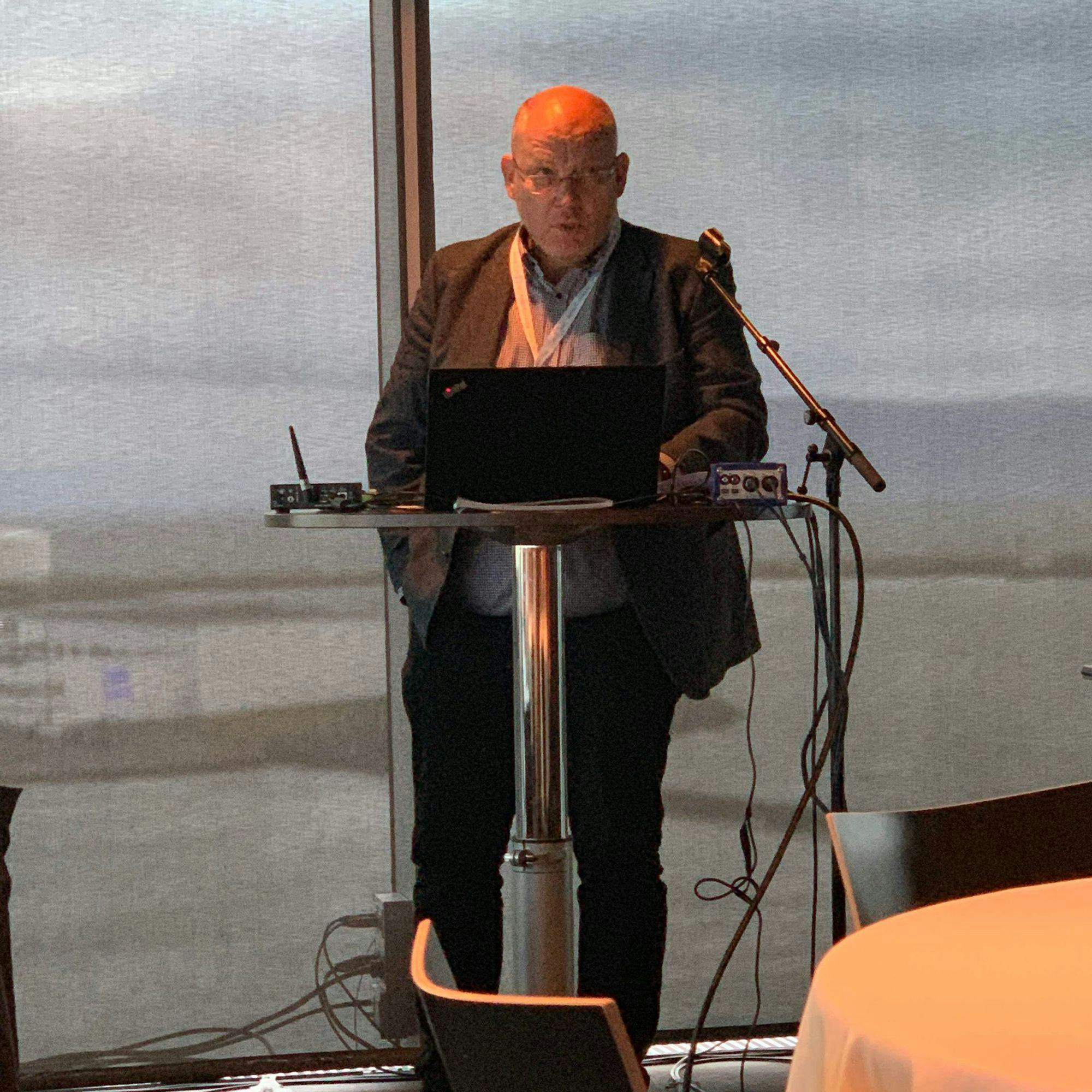 A man speaking into a microphone with a laptop and audio equipment set against a backdrop of large windows overlooking a lake