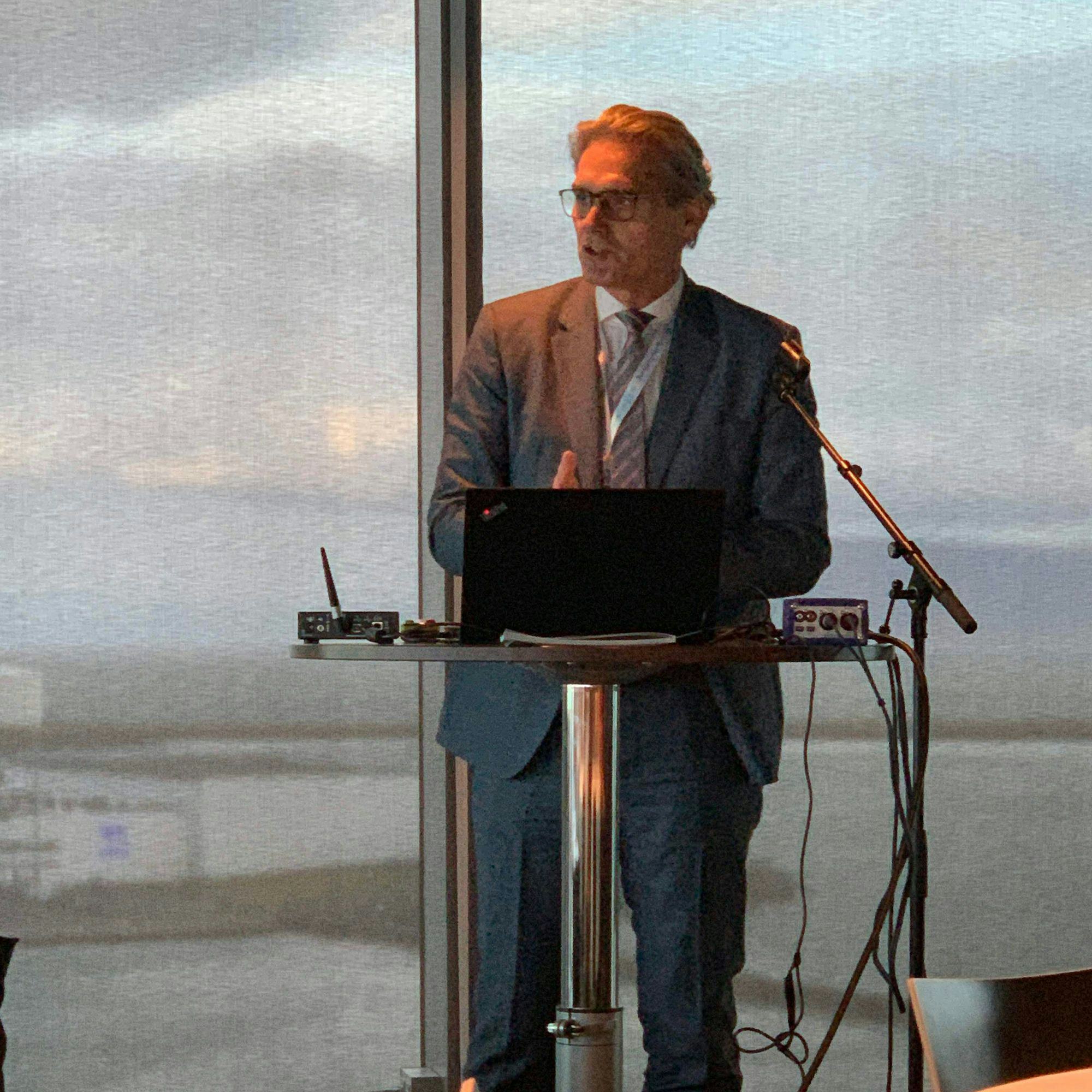 A man speaking into a microphone with a laptop and audio equipment set against a backdrop of large windows overlooking a cloudy sky