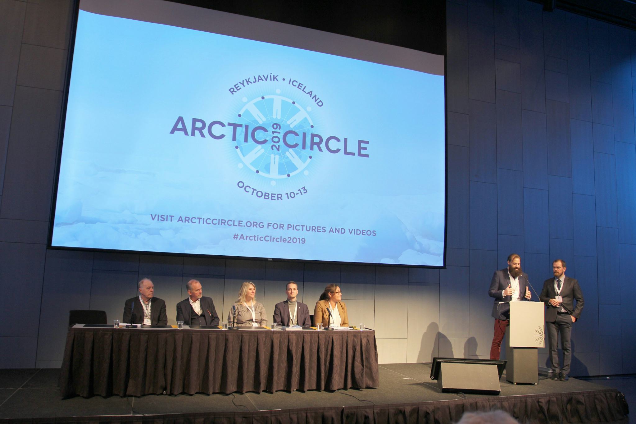 Six individuals seated at a long table and two speakers at a podium, in front of a large screen displaying "ARCTIC CIRCLE"