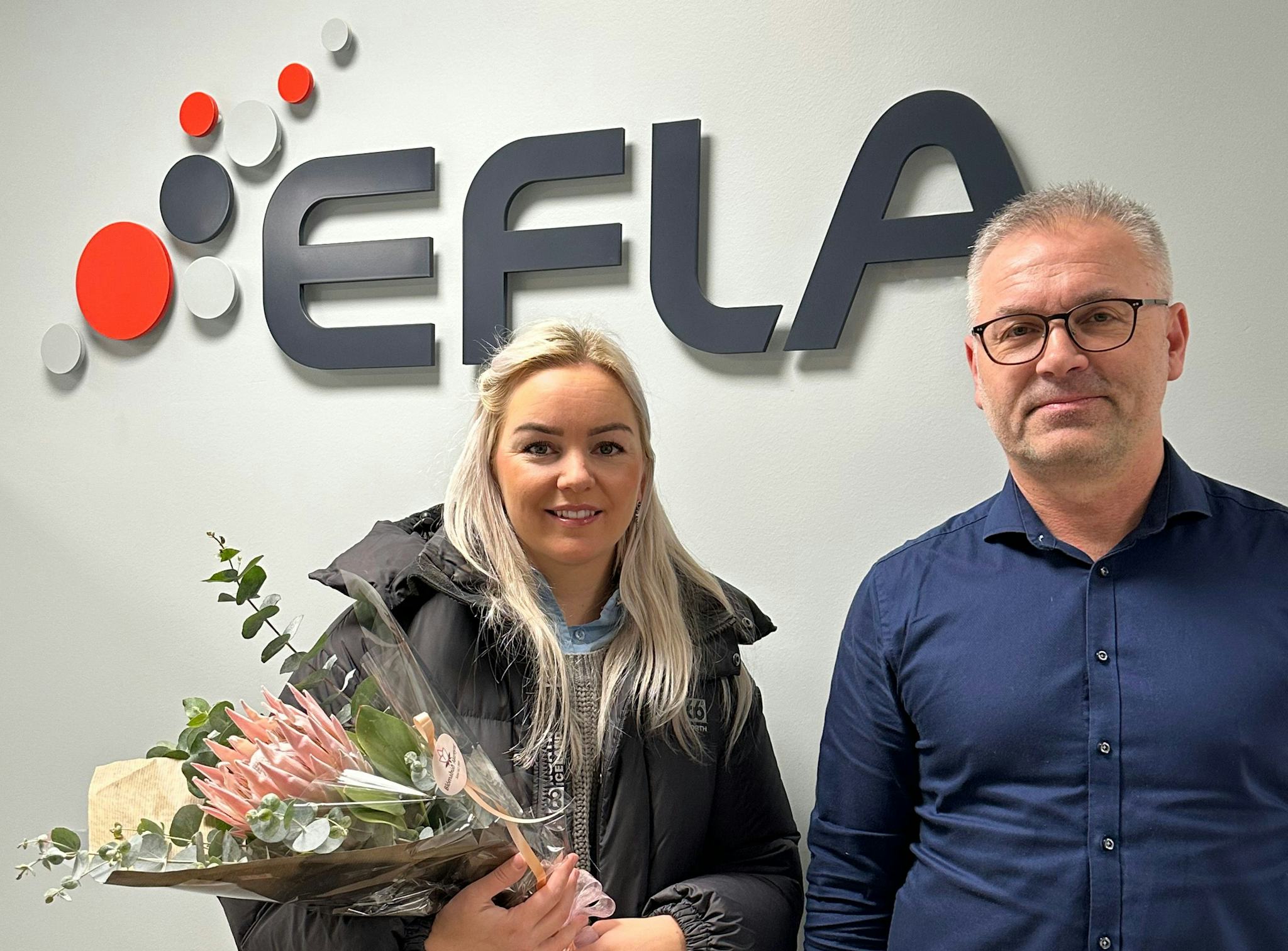 Two individuals standing in front of wall with the EFLA logo, one of them is holding a bouquet of flowers