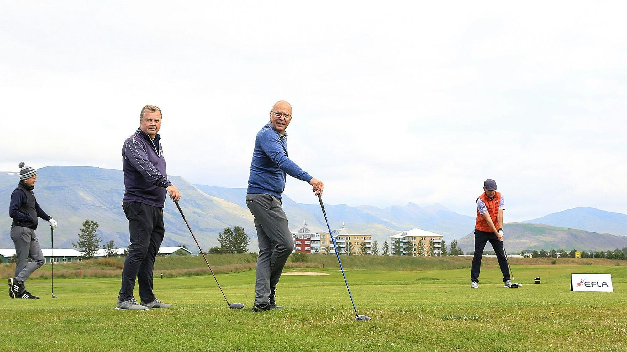 Three men on a golf course, two of them are posing with their clubs and the third one in mid-swing, against a background of mountains and buildings