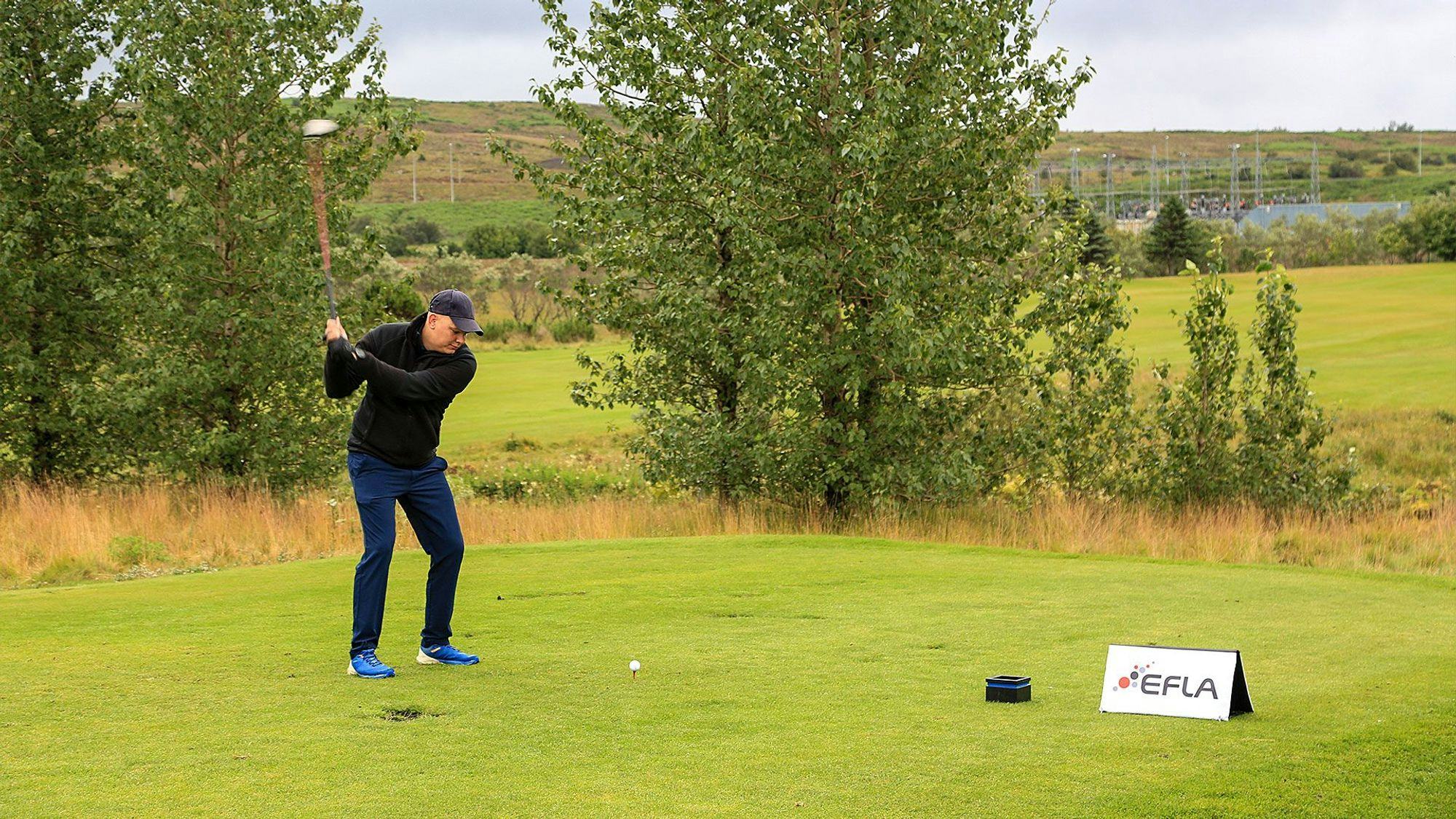 A man in black clothing in mid-swing on a lush golf-course