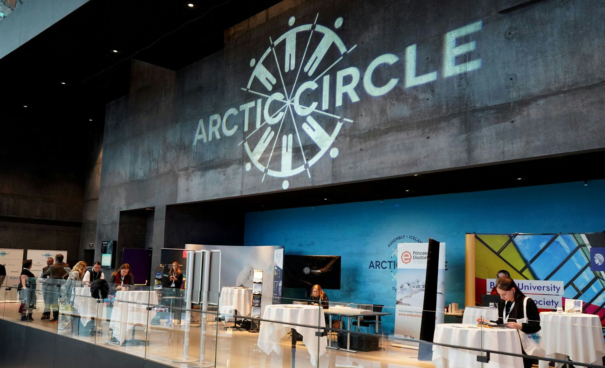 A registration or information area with white cloth covered tables set against a large wall with "ARCTIC CIRCLE" logo displayed